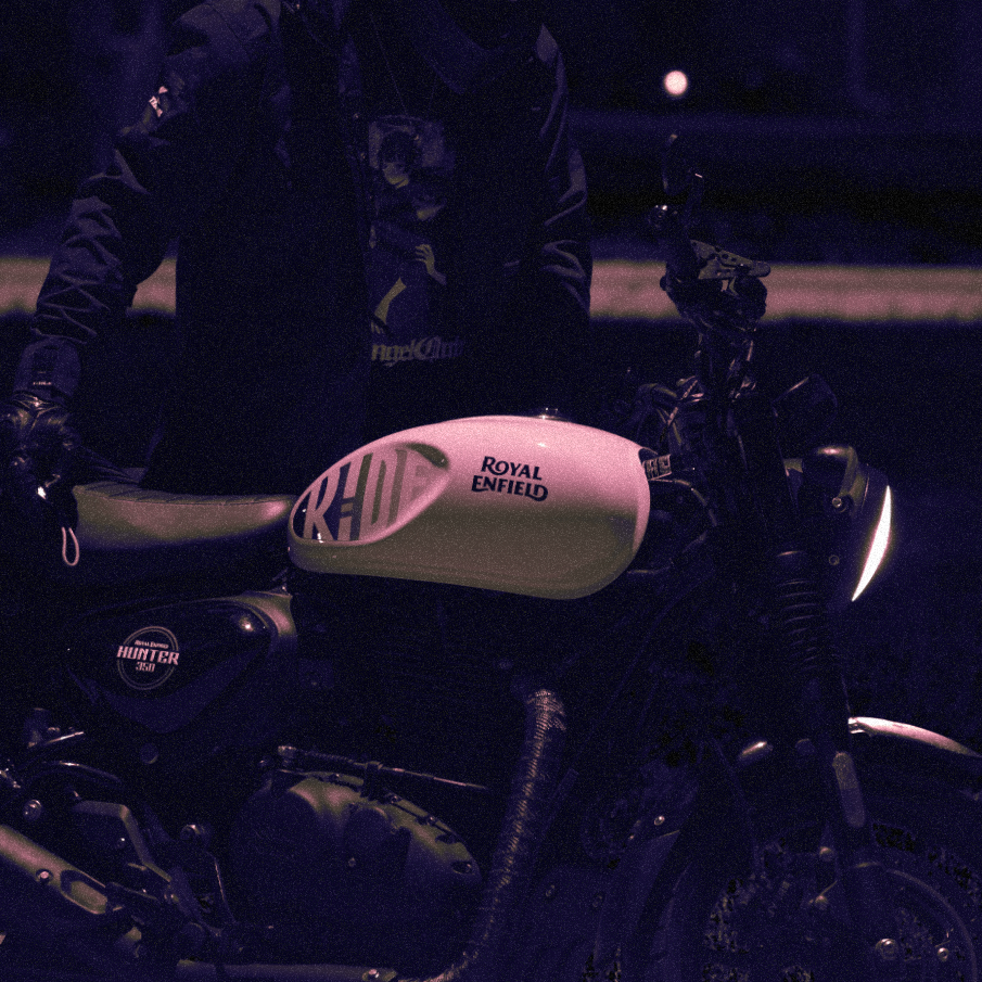 Stylized Photo of a man standing behind his motorcycle. His helment is outside of frame, and the logo of Royal Enfield is prominently displayed on the tank